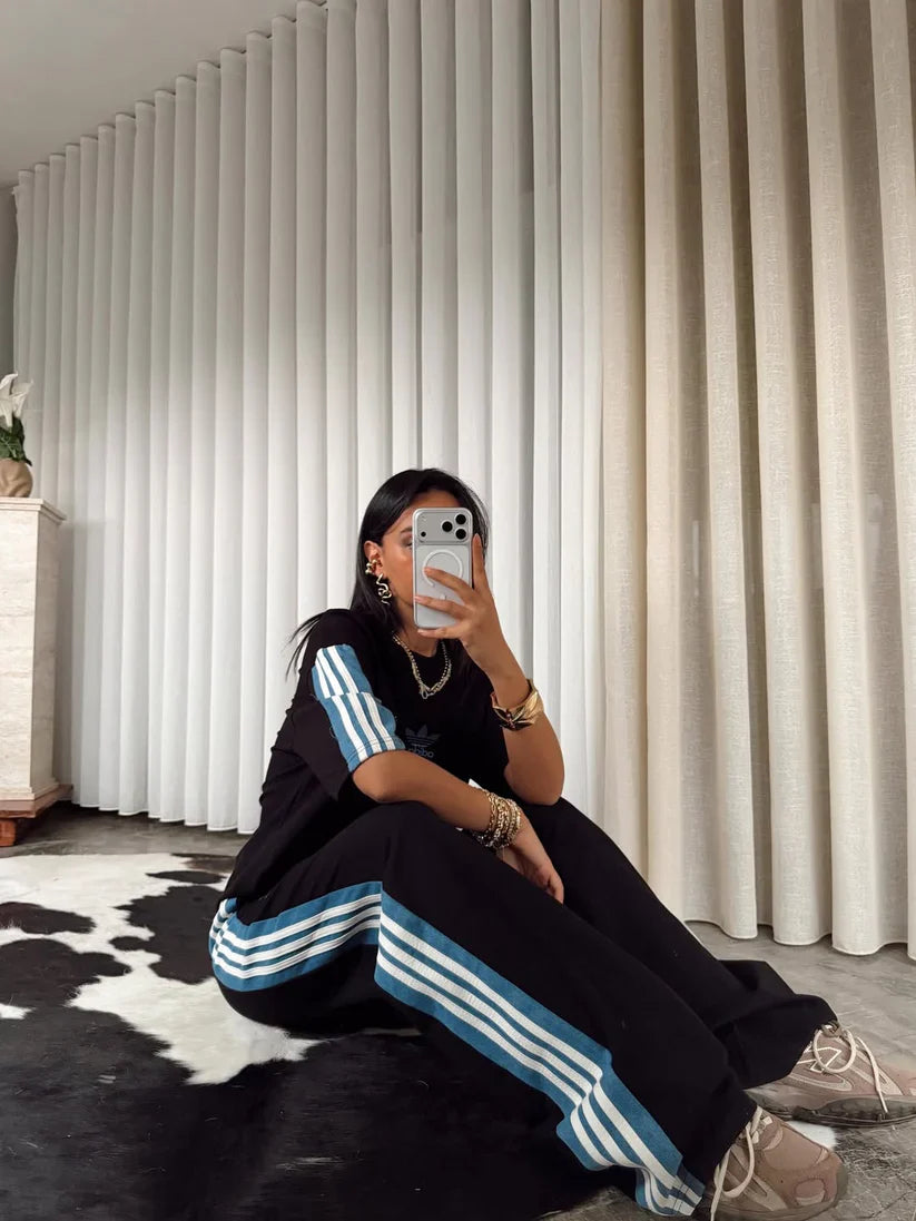 Stylish woman in black oversized co-ord set with striped track pants, sitting indoors and taking a mirror selfie. 01